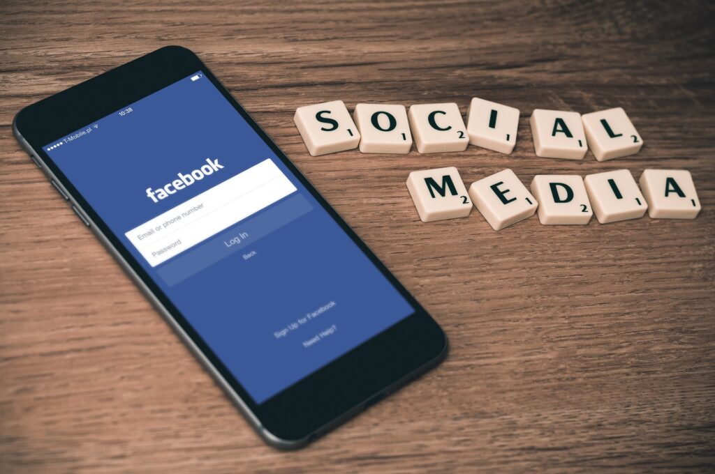 Alt text:
Smartphone displaying Facebook login screen on a wooden table next to tiles spelling “social media,” representing key concepts for Facebook Ads Interview Questions.
